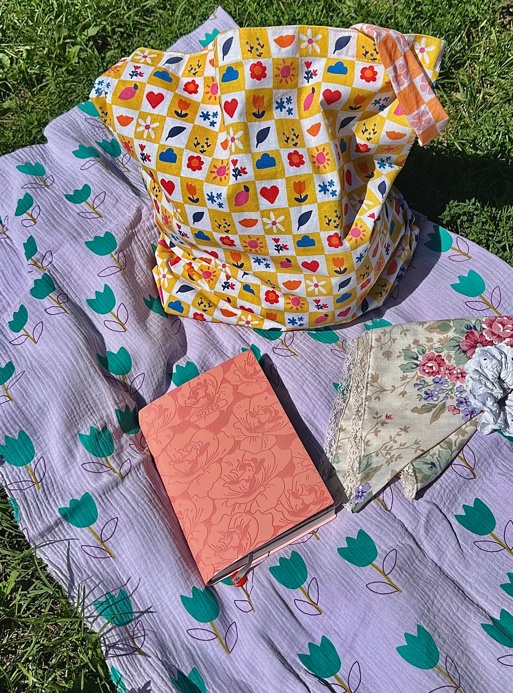 Sac Lisette damier fleuri avec carnet rose et accessoires sur nappe lilas