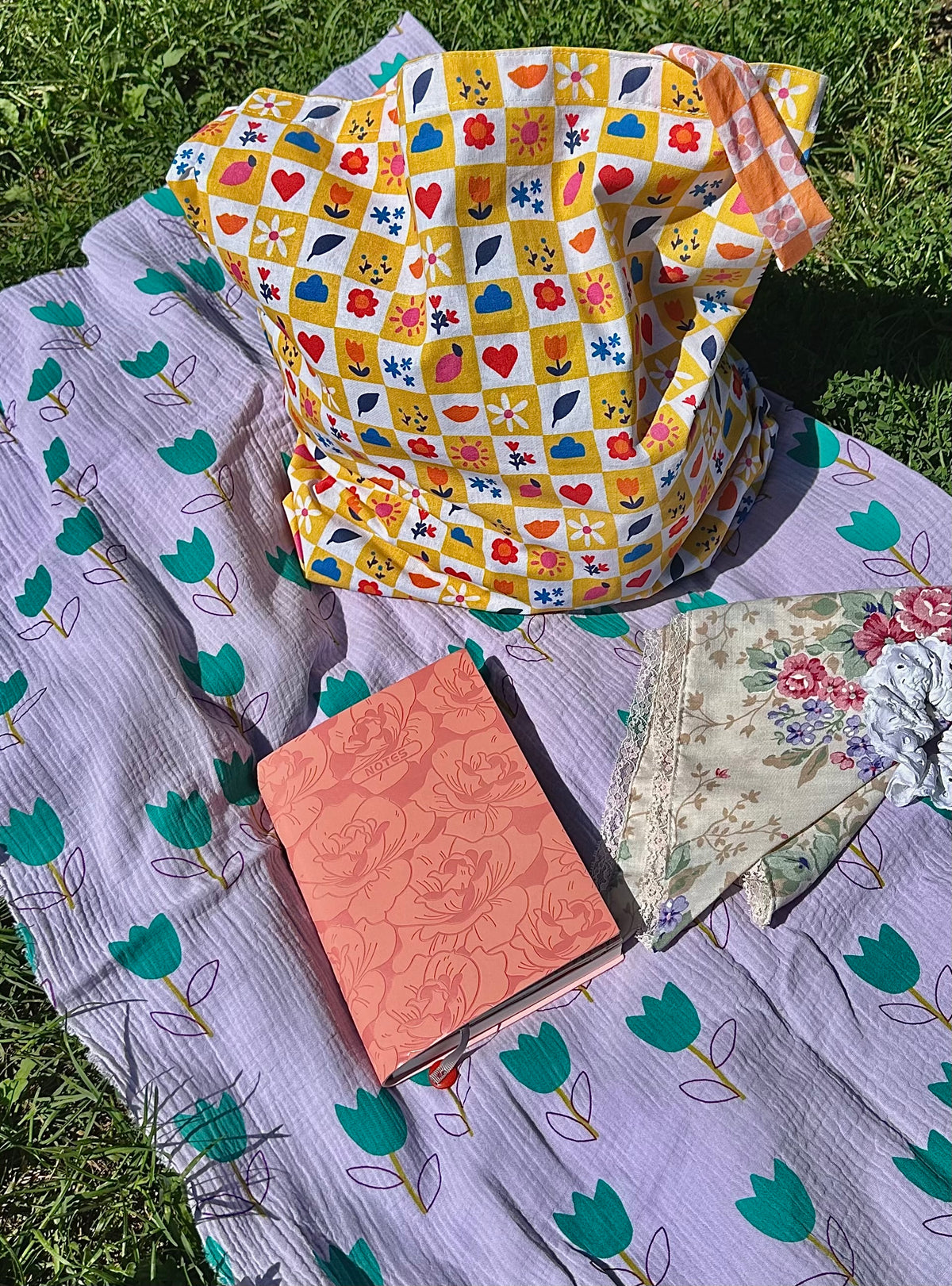 Sac Lisette damier fleuri avec carnet rose et accessoires sur nappe lilas